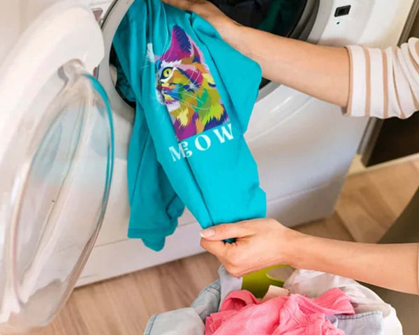Person loading colorful DTF print t-shirts into a washing machine, showcasing DTF print durability tips.